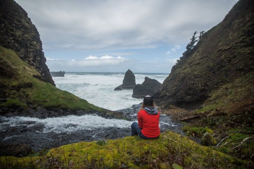 A person in a red shirt sits by a stream, overlooking a rocky coastline and ocean under a cloudy sky.
