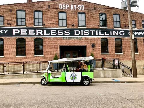 A colorful electric vehicle parked in front of the Peerless Distilling Company building.