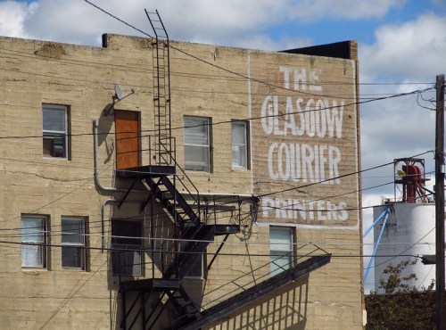 A weathered building with a fire escape and a faded sign reading "The Glasgow Courier Printers" against a cloudy sky.