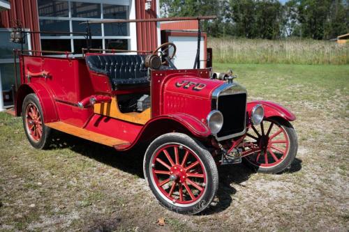 A vintage red car with wooden accents parked on grass near a barn, featuring a black leather interior.