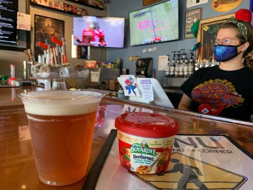 A bar scene featuring a beer in a glass and a cup of Chef Boyardee Beefaroni, with a bartender in the background.