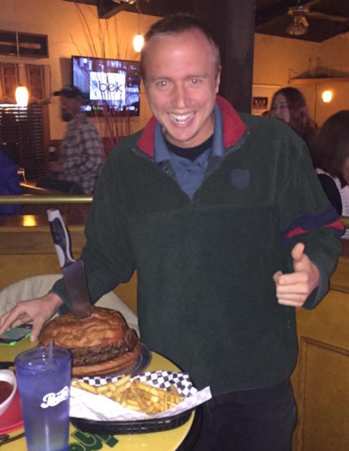 A smiling man in a green sweater holds a large burger with a knife, thumbs up, with fries and a drink on the table.