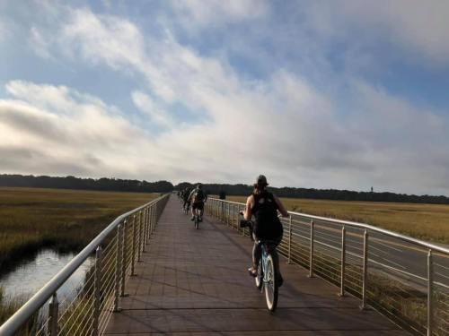 A group of cyclists riding on a wooden boardwalk through a marshy landscape under a cloudy sky.