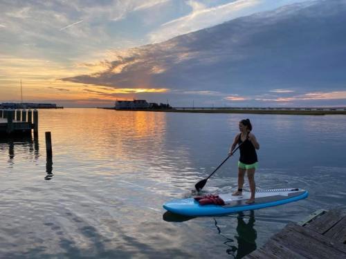 A person paddleboarding on calm water at sunset, with colorful clouds and a distant shoreline in the background.