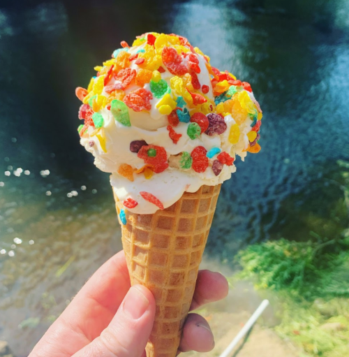 A hand holds a colorful ice cream cone topped with whipped cream and fruity cereal, with a river in the background.