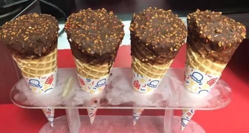 Four chocolate-dipped ice cream cones with sprinkles, displayed on a tray with ice.