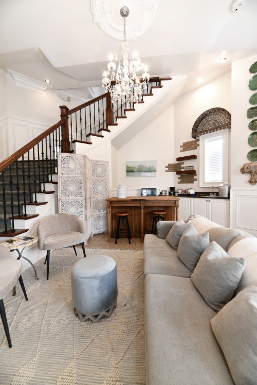 Cozy living room with a staircase, chandelier, gray furniture, and a wooden bar area, featuring decorative wall elements.