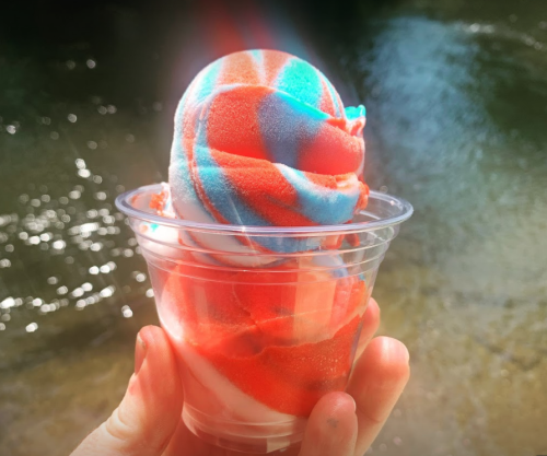 A hand holding a cup of red, white, and blue swirled ice cream against a blurred water background.