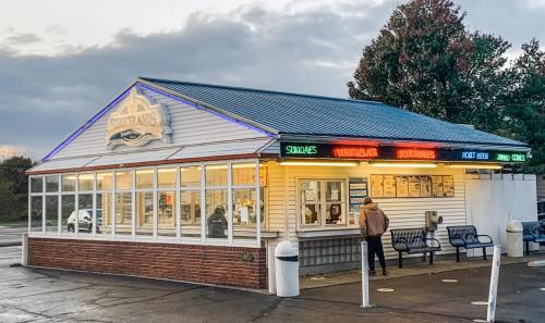 A colorful ice cream stand with a menu, outdoor seating, and a person waiting outside under a cloudy sky.