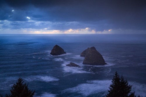 Moody seascape featuring two rocky islands amidst turbulent waves and a cloudy sky at dusk.