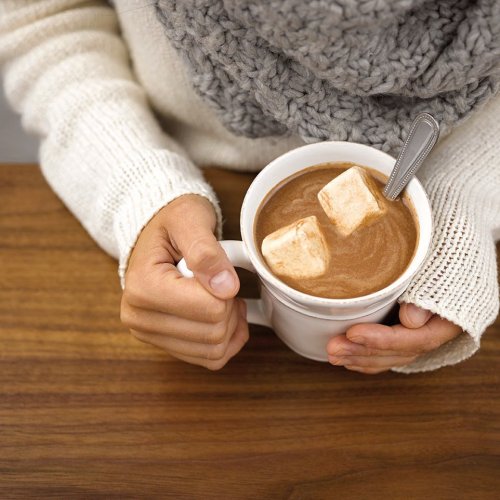 A person holding a mug of hot chocolate topped with marshmallows, wearing a cozy sweater and scarf.