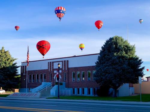 Colorful hot air balloons float above a brick building, with a clear blue sky and trees in the background.