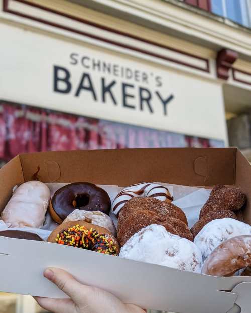 A hand holding a box of assorted donuts in front of Schneider's Bakery sign.