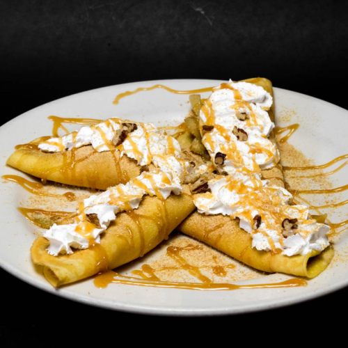 A plate of crepes topped with whipped cream, caramel drizzle, and crushed nuts, served on a dark background.