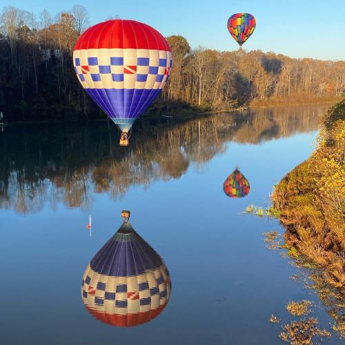 Two hot air balloons float above a calm river, reflecting their colors in the water, surrounded by autumn trees.