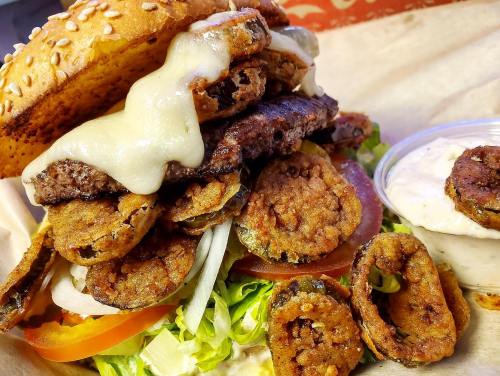 A towering burger with layers of beef, fried pickles, cheese, lettuce, and tomato, served with a side of dipping sauce.