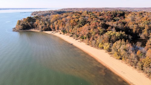 Aerial view of a sandy beach bordered by colorful autumn trees along a calm lake.