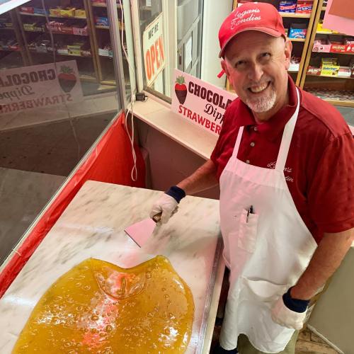 A smiling man in a red cap and apron prepares a large sheet of yellow candy in a shop with a bright, welcoming atmosphere.