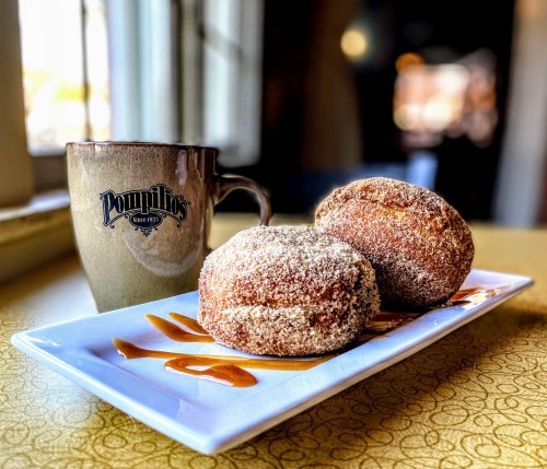 A plate with two cinnamon sugar donuts drizzled with caramel, next to a coffee mug on a table.