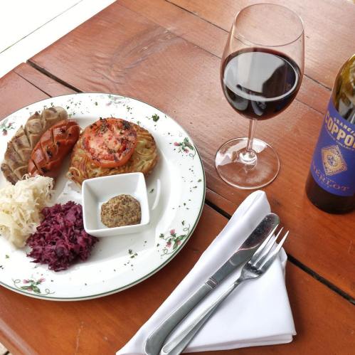 A plate with pasta, sausage, sauerkraut, and mustard, accompanied by a glass of red wine and a bottle on a wooden table.