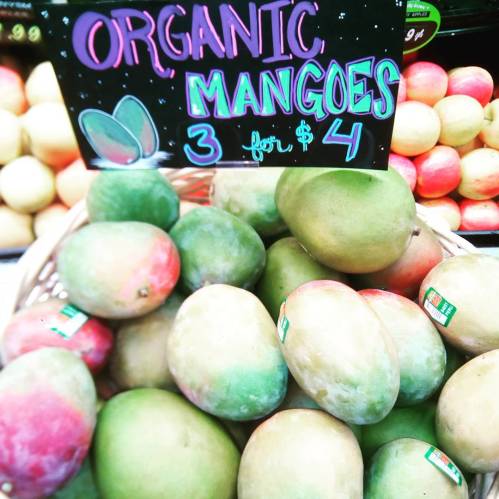 A basket of organic mangoes with a sign reading "Organic Mangoes 3 for $4" in a colorful market display.