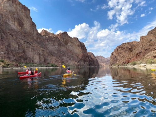 Two kayakers paddle on a calm river surrounded by towering rocky cliffs and a blue sky with scattered clouds.