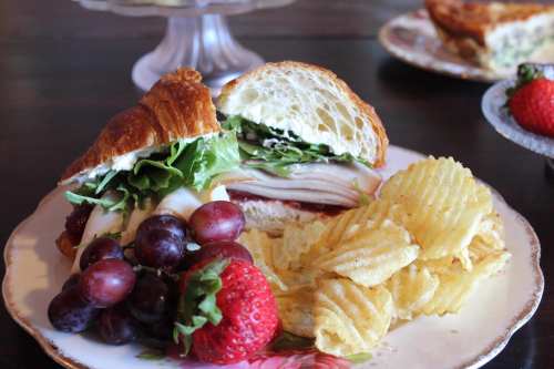 A croissant sandwich with turkey, lettuce, and mayo, served with grapes, strawberries, and potato chips on a floral plate.