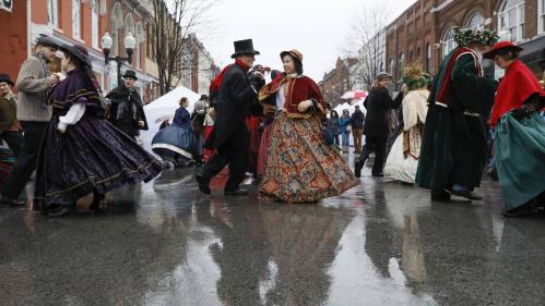 A group of people in Victorian costumes dance on a rainy street, reflecting colorful outfits in puddles.