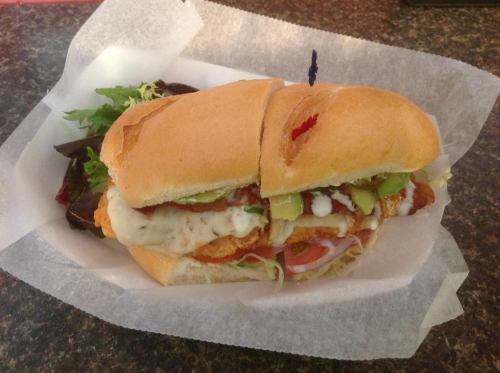 A large sandwich with fried chicken, lettuce, tomatoes, pickles, and sauce, served with a side of greens.