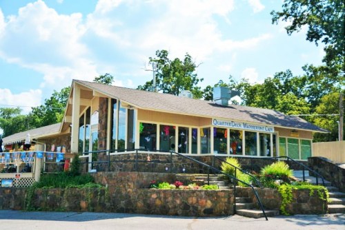 A charming stone building with large windows, featuring a sign for "QuarterDuck Waterfront Cafe" and surrounded by greenery.