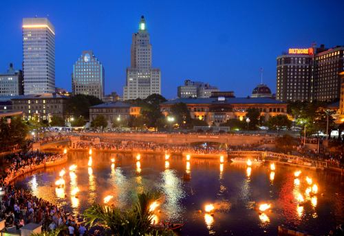 A vibrant city skyline at dusk, with flames reflecting on a river and a crowd gathered along the water's edge.