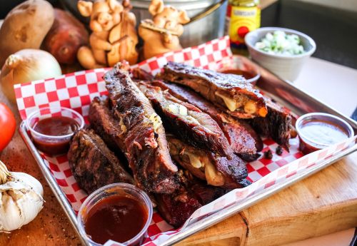 A platter of smoked ribs with barbecue sauce, garnished with vegetables, on a checkered paper-lined tray.