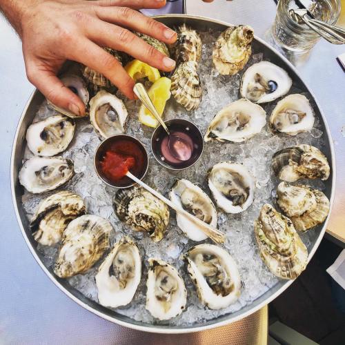 A metal platter filled with ice and fresh oysters, accompanied by lemon wedges and dipping sauces.