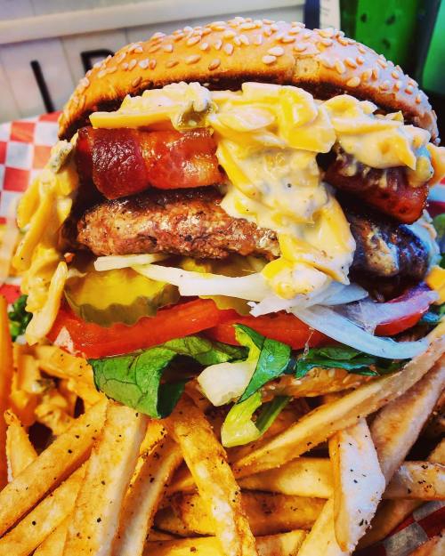A towering burger with bacon, cheese, lettuce, tomato, and pickles, served with a side of crispy French fries.