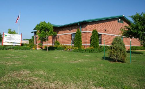 A brick building with a green roof, surrounded by grass and trees, featuring a sign and an American flag.