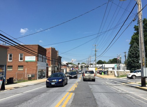 A view of a small town street with cars, buildings, and power lines under a clear blue sky.