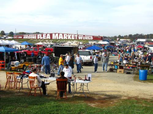 A bustling flea market with vendors, shoppers, and colorful tents under a cloudy sky.