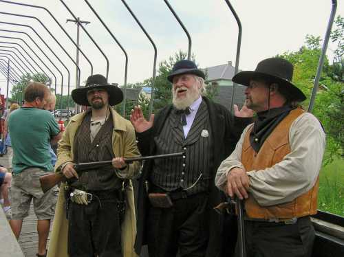 Three men in period costumes, one with a rifle, stand in a train car, engaging with a crowd in a lively setting.