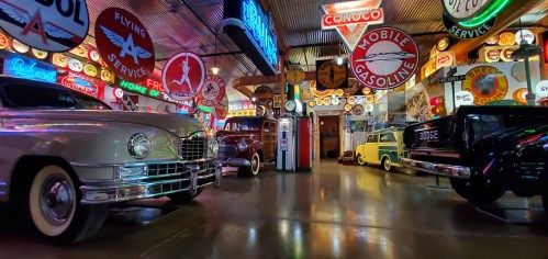 A vintage car museum filled with classic cars and colorful gas station signs, illuminated by neon lights.