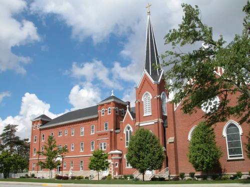 A red brick church with a tall steeple and cross, surrounded by trees under a partly cloudy sky.