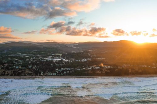 Aerial view of a coastline at sunset, with waves crashing and hills in the background. Soft clouds in the sky.