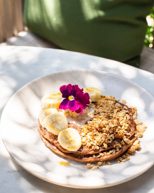 A plate of pancakes topped with banana slices, granola, and a purple flower, set on a light table.