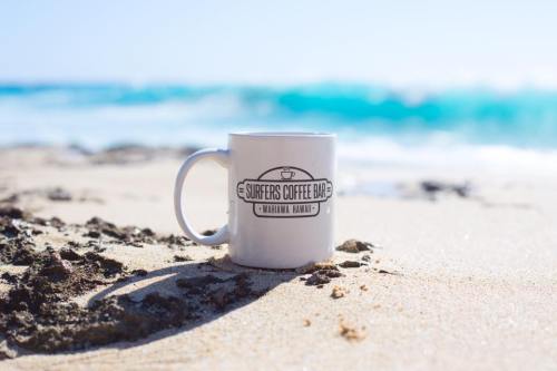A white coffee mug with "Surfers Coffee Bar" logo sits on sandy beach, with ocean waves in the background.