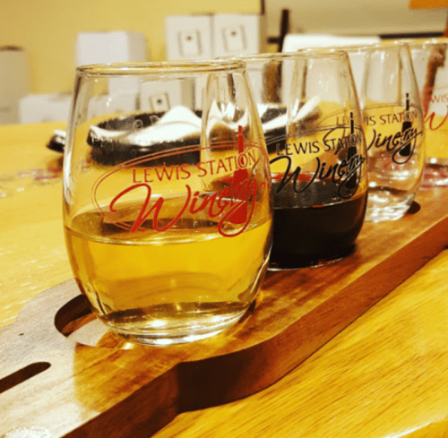 A wooden flight tray holds three wine glasses: one with white wine and two with red wine, all branded with "Lewis Station."