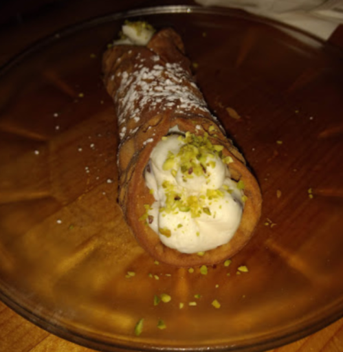 A chocolate cannoli filled with creamy filling and topped with crushed pistachios, served on a plate.