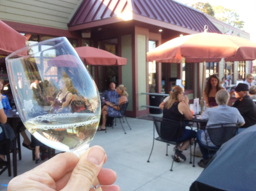 A hand holding a glass of white wine in front of a lively outdoor dining area with people seated under umbrellas.