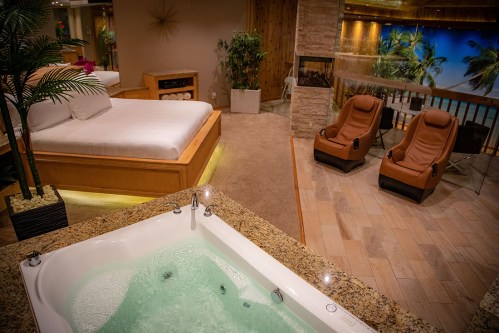 A luxurious spa room featuring a bed, jacuzzi, and two reclining chairs with a tropical backdrop.