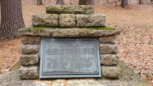 Stone monument with a plaque marking the site of "Old Helen," surrounded by trees in a wooded area.