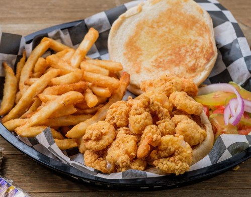 A basket of fried shrimp, seasoned fries, a toasted bun, and fresh vegetables on a checkered paper lining.