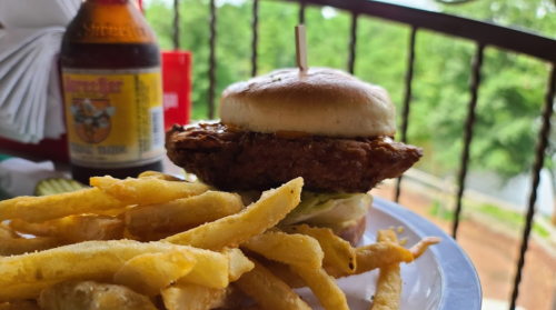 A close-up of a fried chicken sandwich with lettuce on a plate, accompanied by golden fries and a drink in the background.